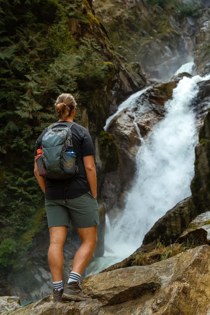 A hiker stands on a rocky ledge facing a powerful waterfall surrounded by steep cliffs and dense evergreen forest. He wears a black shirt, green shorts, and a large backpack with water bottles tucked into side pockets.