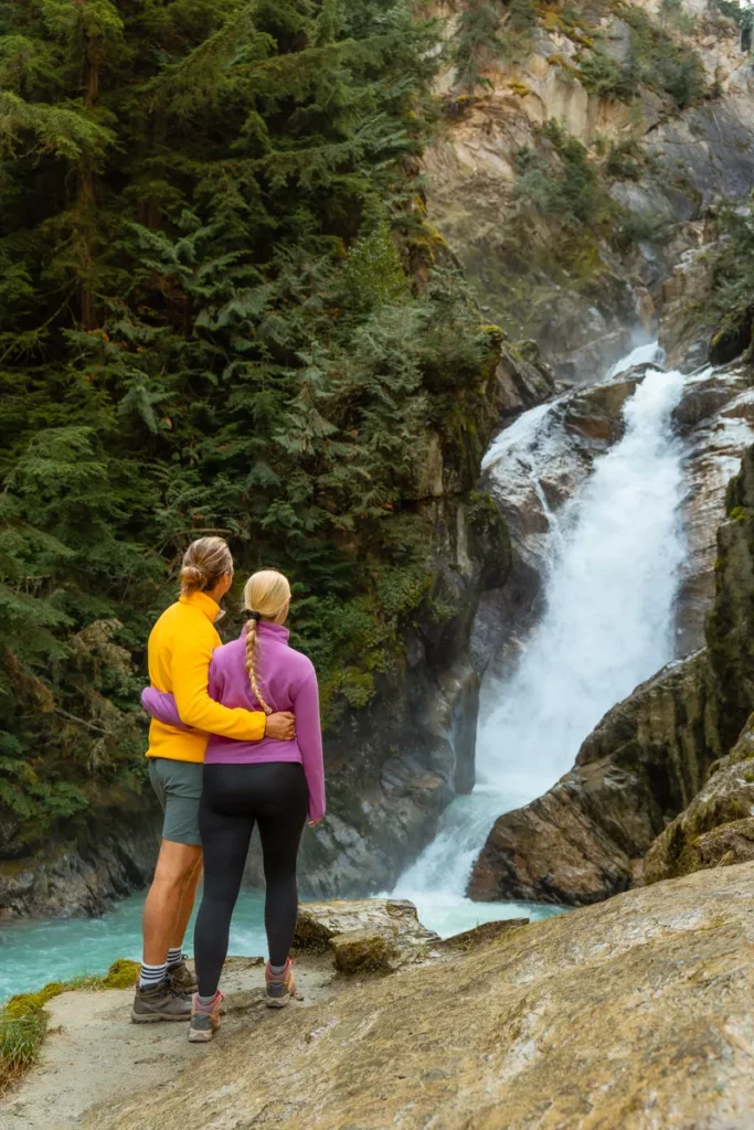 A couple stands arm in arm on a rocky ledge, looking at a waterfall cascading through a forest. The man wears a bright yellow sweater and the woman a pink one, both dressed in hiking gear, surrounded by lush evergreen trees and turquoise water below.