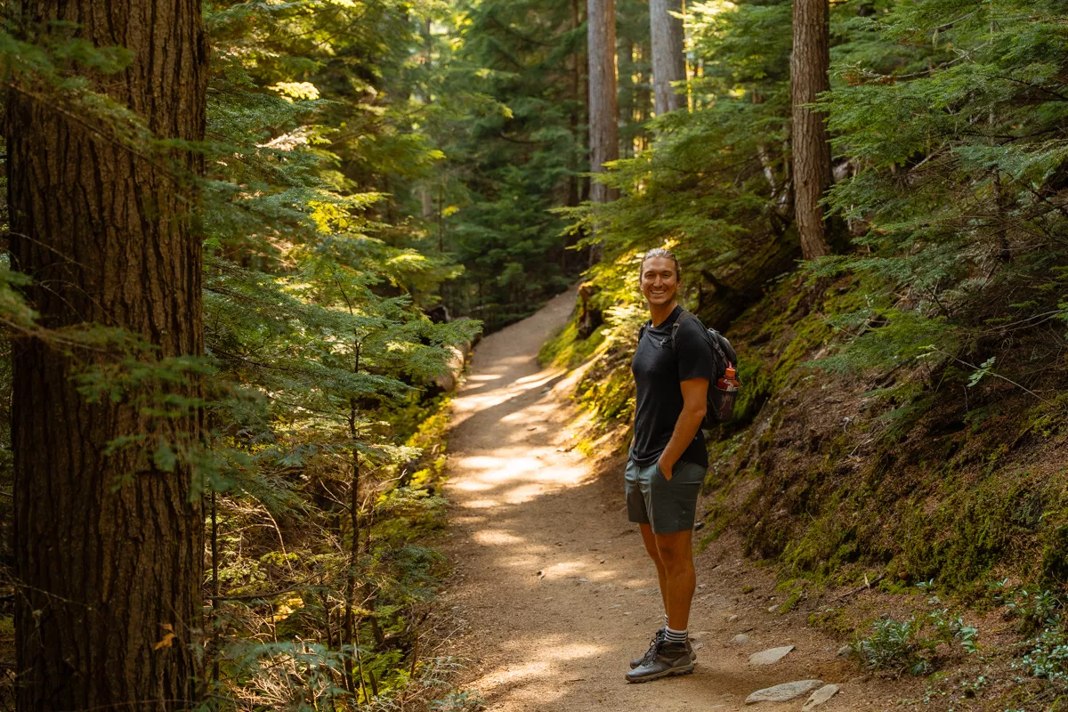A smiling hiker stands on a sun-dappled forest trail, surrounded by tall trees. He wears a black shirt, green shorts, and hiking boots, with a backpack carrying a water bottle.