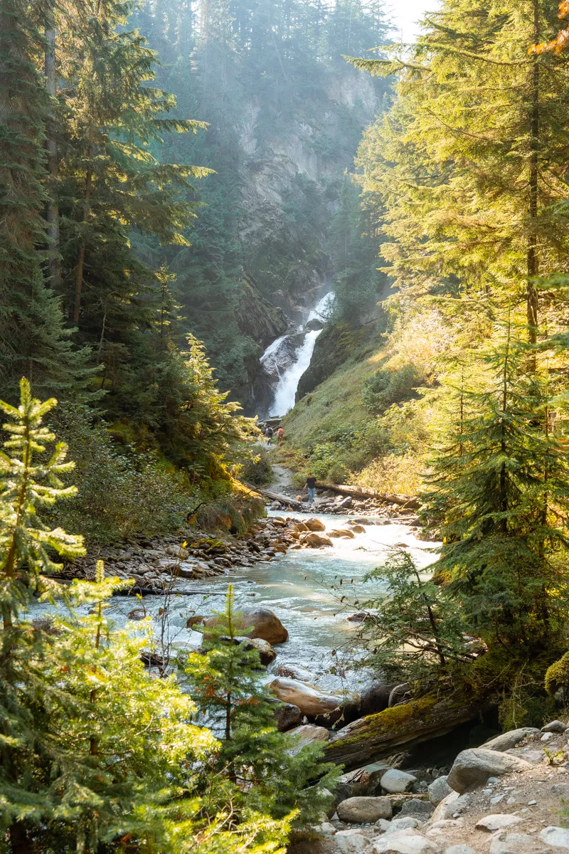 A scenic view of a cascading waterfall in a lush forest, with a clear stream flowing over rocks and hikers visible on the trail leading toward the falls.