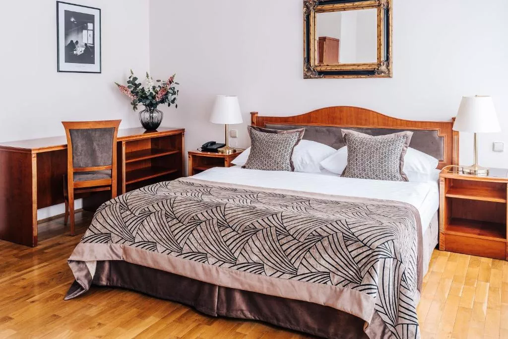 A classic hotel room with a wooden double bed framed by matching nightstands and a desk set, featuring neutral bedding with a patterned throw and three textured accent pillows. The space includes warm wood flooring, two white lamps, a framed mirror above the bed, and a vase of flowers adding a touch of decor.