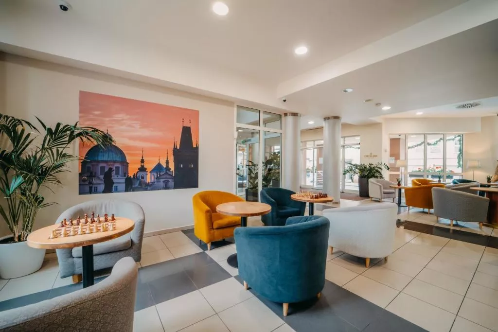 A bright and modern hotel lounge area with cozy armchairs in various colors arranged around small wooden tables, two of which have chess boards set up. A large photo of Prague’s skyline at sunset decorates the wall.