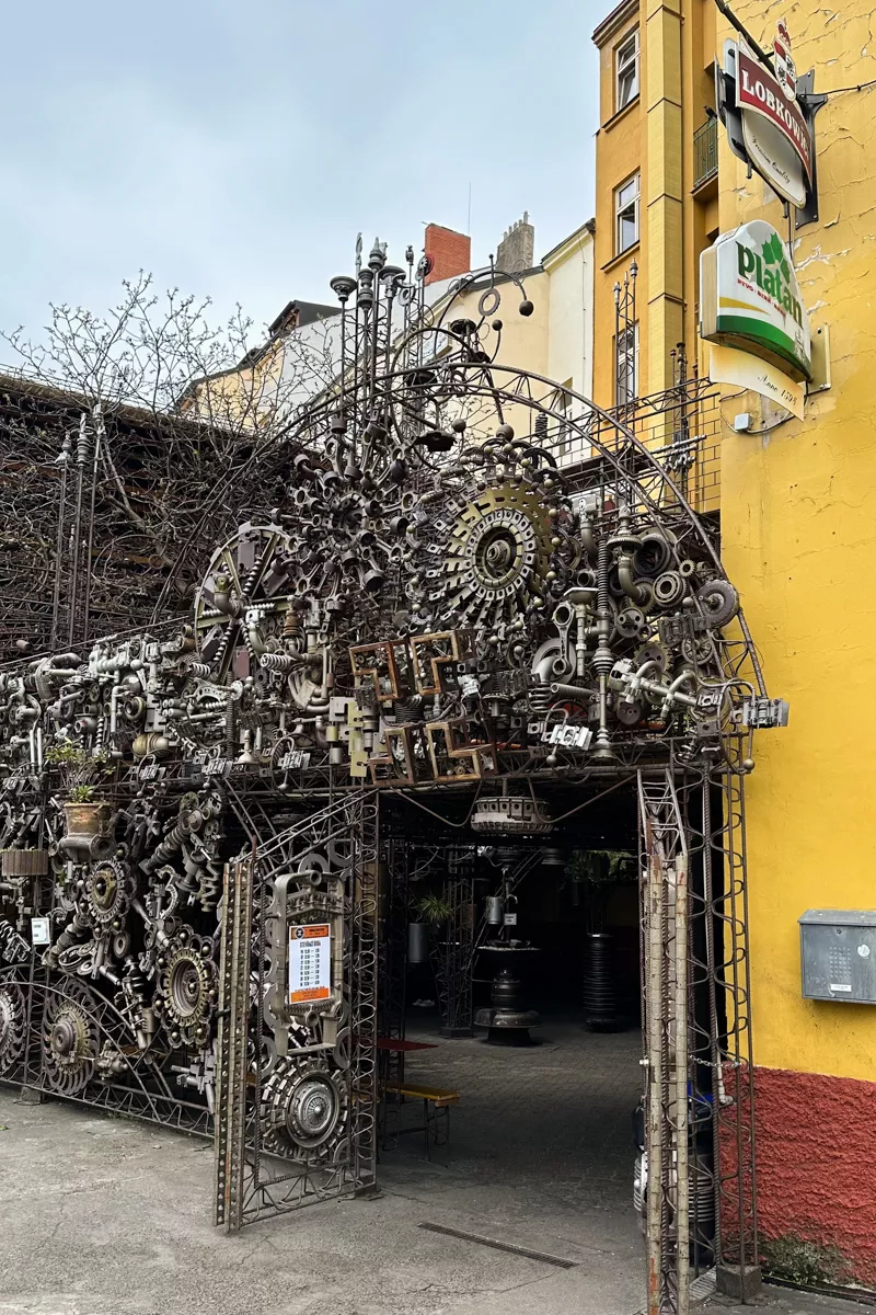 An intricate metal gate entrance to a courtyard bar in Prague, made entirely of gears, pipes, and mechanical parts, forming a detailed industrial art installation. The gate is set against a bright yellow building with signage for “Lobkowicz” and “Platan” beers.