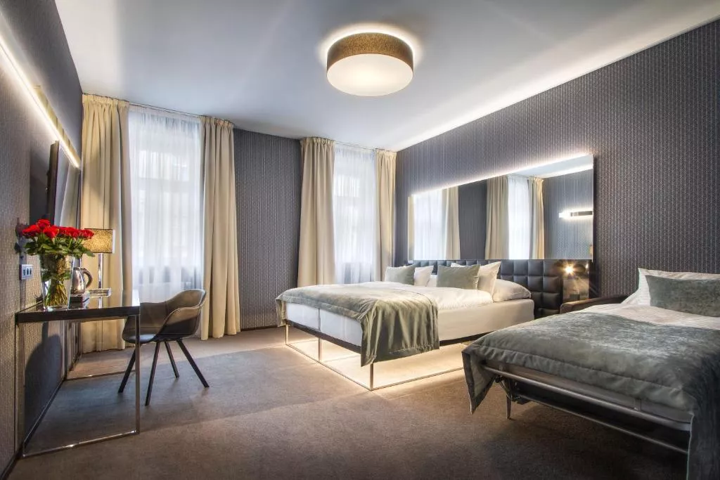 A modern and spacious hotel room with a double bed and a single bed, both featuring soft gray-green bedding. The room is styled with textured dark wallpaper, a large illuminated mirror, cream curtains over tall windows, and a nice desk setup.
