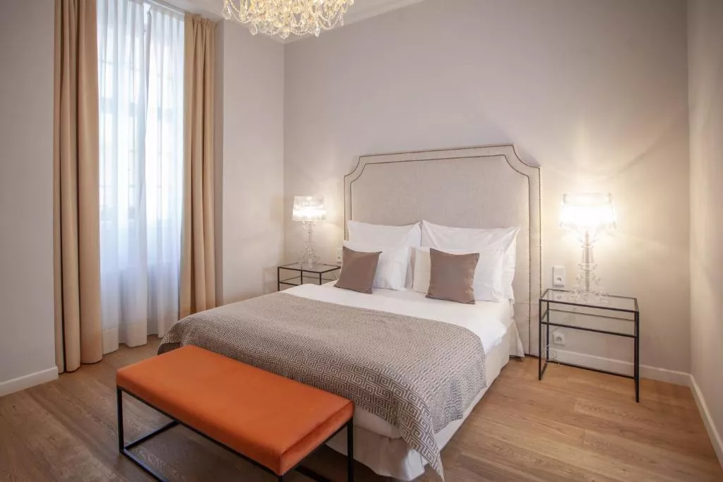 A bright and elegant hotel room featuring a large upholstered headboard, neutral-toned bedding with three decorative pillows, and a soft taupe blanket.