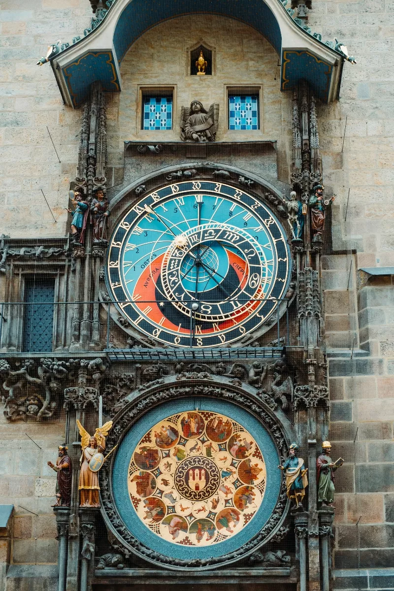 A richly detailed image of the Prague Astronomical Clock, featuring its two main dials. The upper dial displays astronomical and zodiac signs in a vibrant blue and gold color scheme, while the lower dial shows a calendar with intricate illustrations of the months and medallions representing the zodiac. Statues of saints and symbolic figures frame the clock’s Gothic structure.