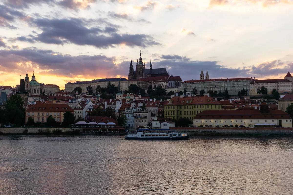 A serene evening view of Prague Castle and St. Vitus Cathedral silhouetted against a dramatic sunset sky, with golden and purple hues peeking through scattered clouds. Below, a boat glides along the Vltava River, passing charming red-roofed buildings lining the riverbank.