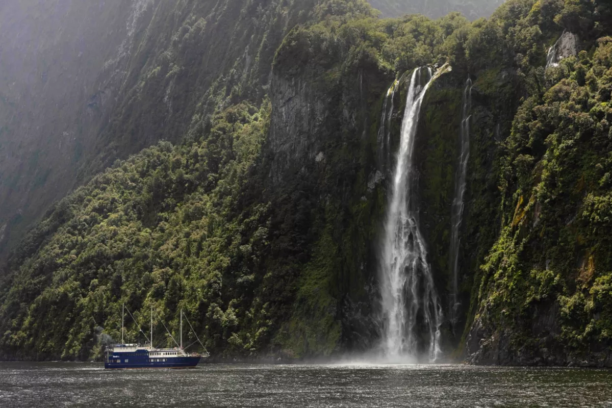 A tall, narrow waterfall cascades dramatically down a moss-covered cliffside into a misty fjord, with a blue cruise boat passing nearby along the forested shoreline. Sunlight filters through, highlighting the lush greenery and the spray at the waterfall's base.