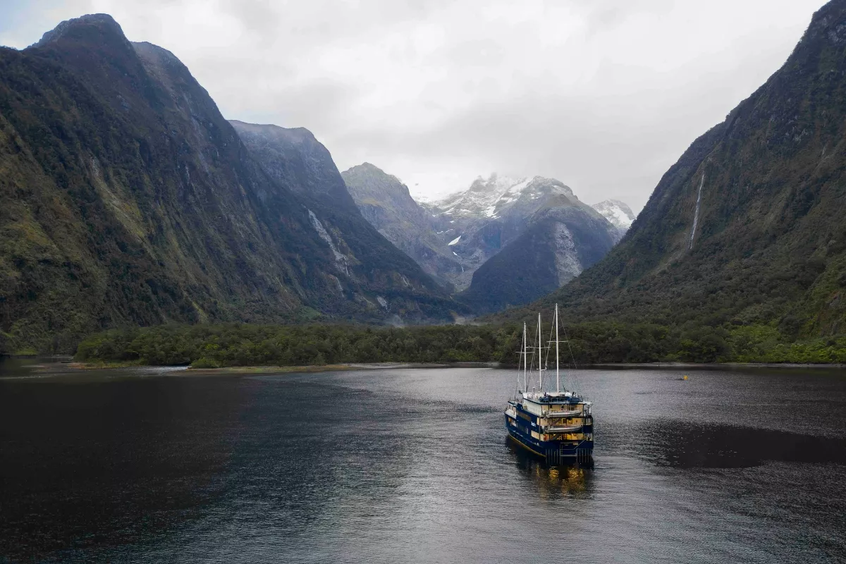 A blue cruise boat floats in a still fjord in Milford, surrounded by steep, forested mountains with snow-capped peaks in the background under a cloudy sky.