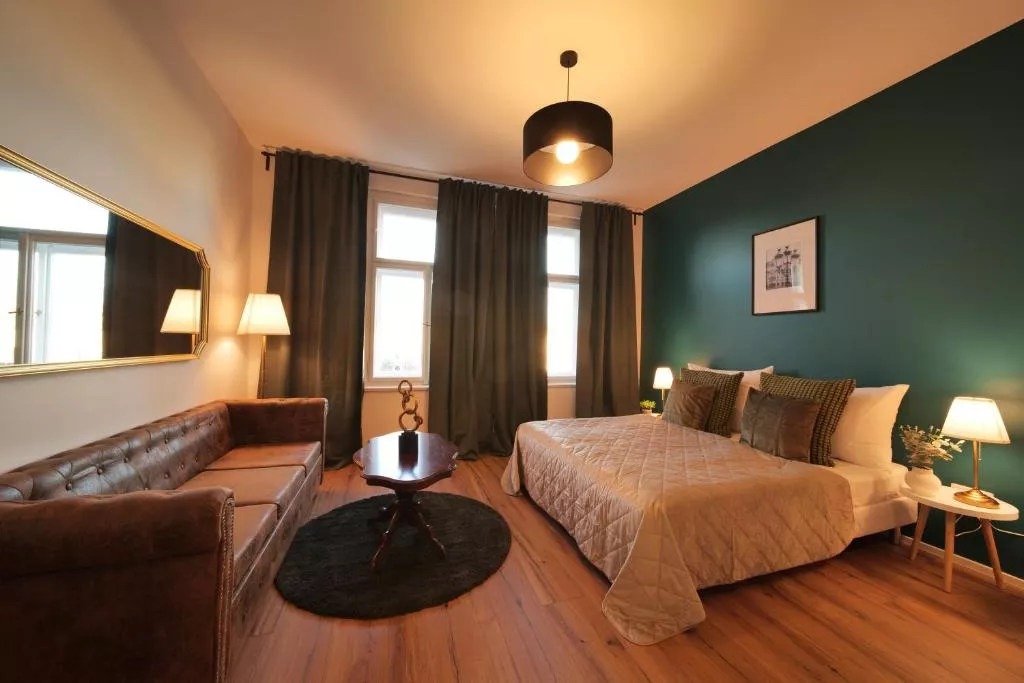 A cozy and stylish bedroom with a large bed dressed in a beige quilt, set against a dark teal accent wall. The room features warm lighting, a tufted leather sofa, dark curtains over tall windows, a large gold-framed mirror, and a round wooden coffee table on a dark rug.