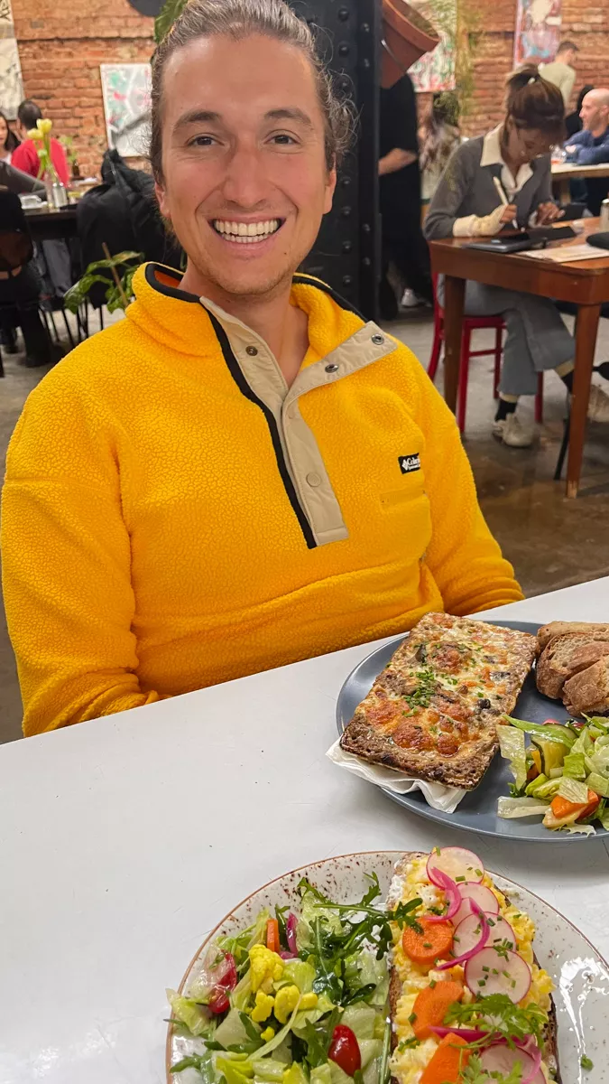 A smiling man in a bright yellow fleece sitting at a table in a cozy restaurant, with a plate of food in front of them featuring toasted bread topped with melted cheese, a side salad, and slices of crusty bread. The background shows other diners and warm, rustic decor with exposed brick walls.