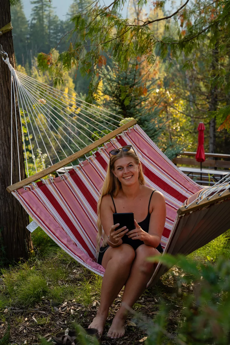 A smiling woman relaxes barefoot on a red-striped hammock strung between trees, holding an e-reader and surrounded by dappled sunlight and forest greenery.