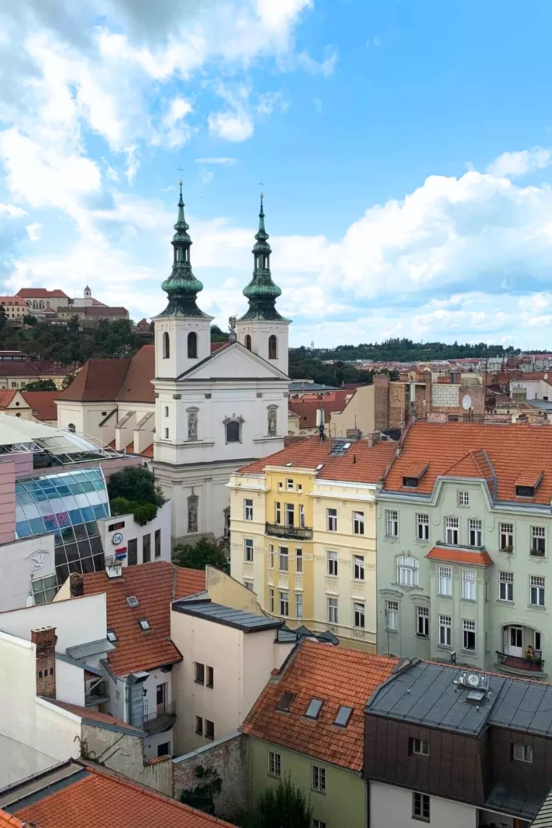 Aerial view over the rooftops of Brno, Czech Republic, featuring the white facade and green spires of the Church of St. Thomas, surrounded by colorful historic buildings and modern glass architecture.