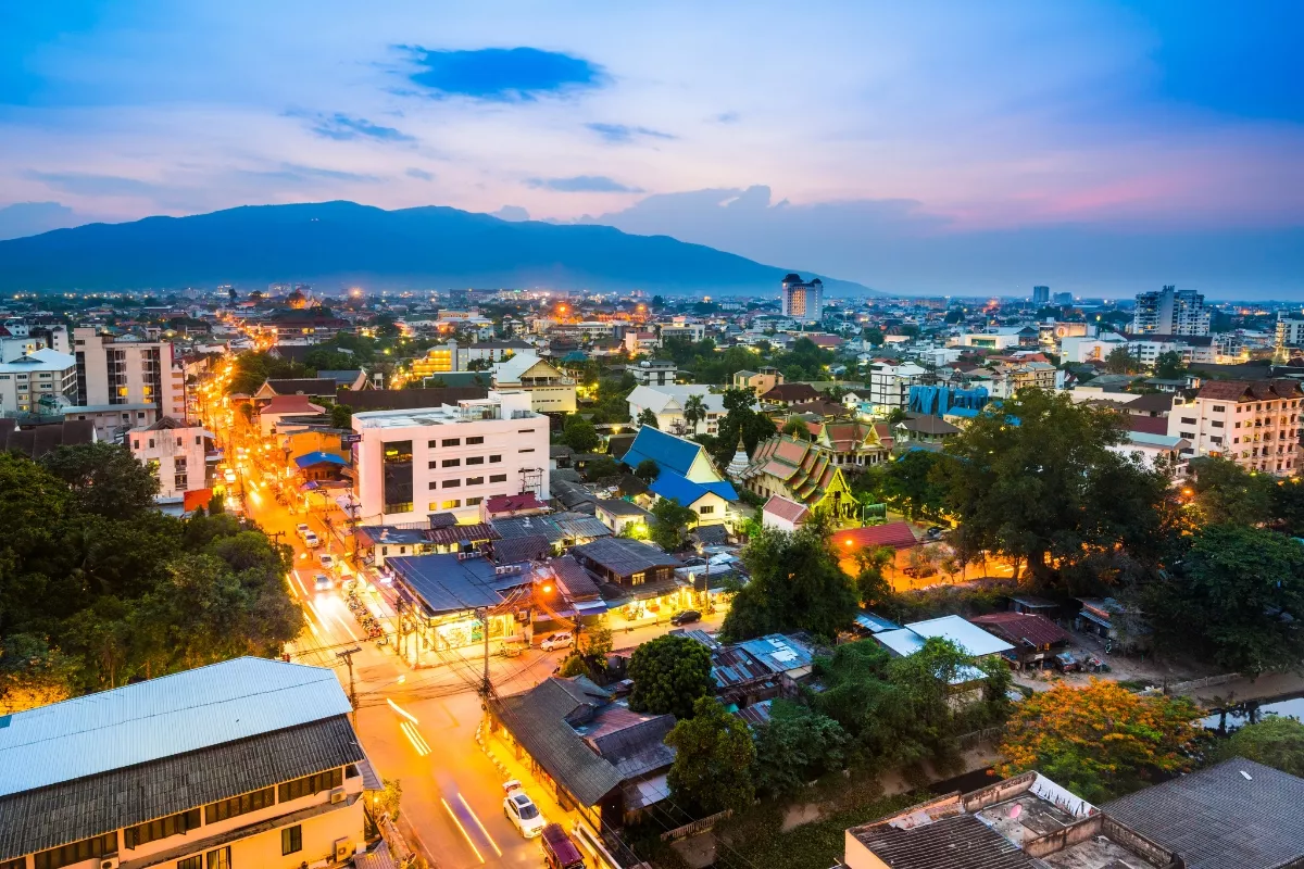 This image shows a vibrant evening cityscape of Chiang Mai, Thailand, with golden streetlights lining the roads and a backdrop of lush mountains under a twilight sky.