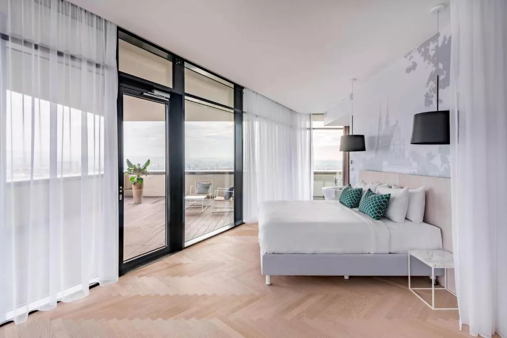This image shows a modern, airy bedroom with floor-to-ceiling windows that open onto a spacious balcony. The room features a minimalist design with soft wood flooring, white curtains, a large bed with white linens and green-accented pillows. The balcony includes a potted plant and seating area, offering panoramic views of the city below.