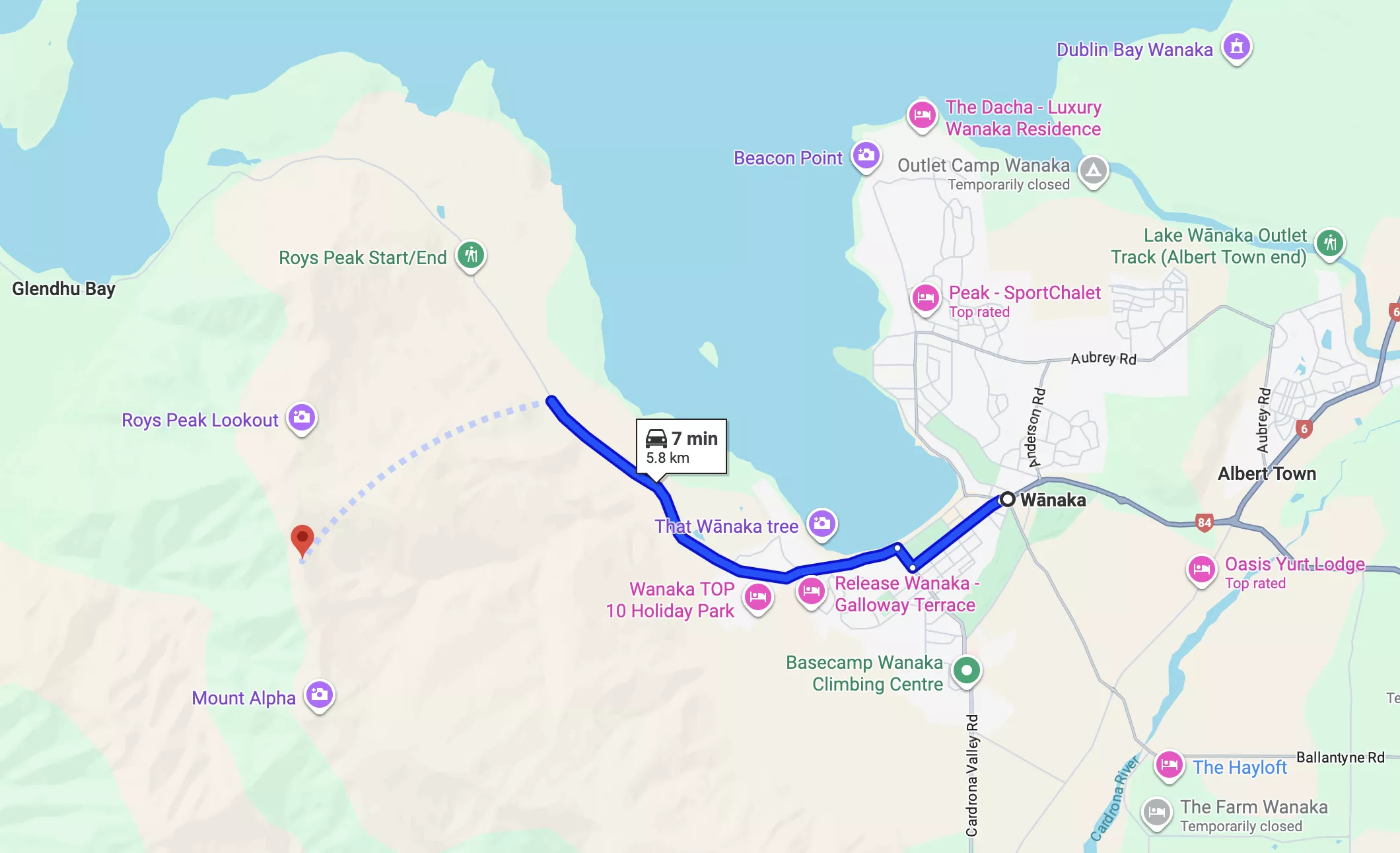 A map showing the driving route from central Wanaka to the Roys Peak trailhead in New Zealand, with a highlighted 5.8 km path taking approximately 7 minutes by car. Key landmarks like That Wanaka Tree, Roys Peak Lookout, and Mount Alpha are also marked along the way.