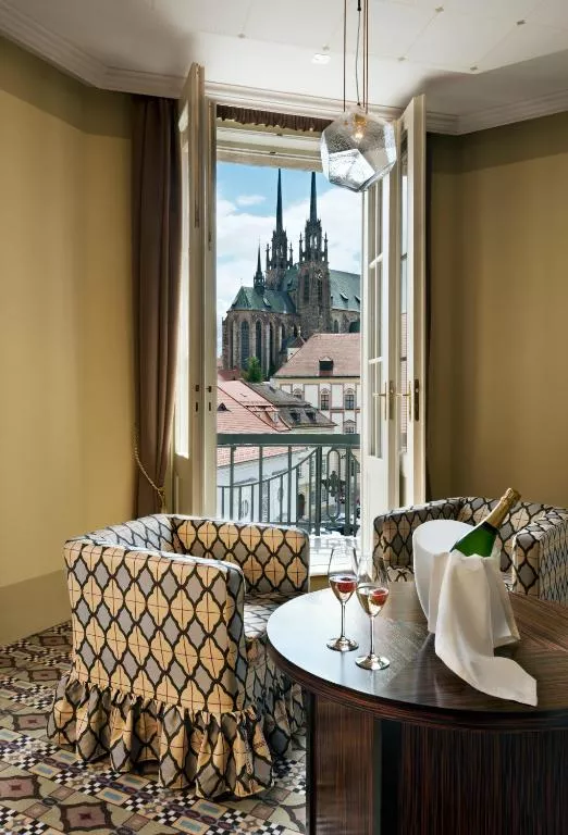 A cozy seating area inside a hotel room with a small round table set for two, featuring a champagne bottle in an ice bucket and glasses of rosé. French doors open onto a balcony with a striking view of the Cathedral of St. Peter and Paul in Brno, Czech Republic.