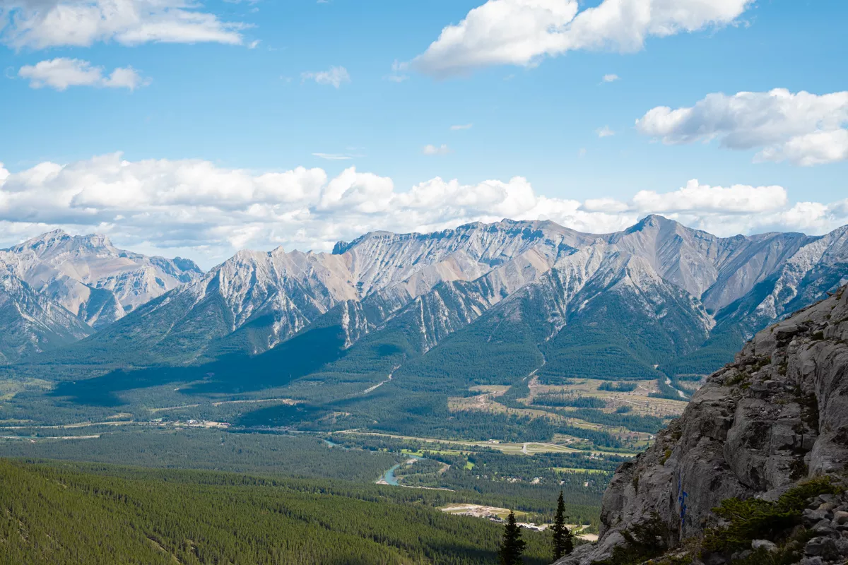 Expansive view from Ha Ling Peak of the Fairholme Range, with its sharply ridged mountains rising beyond a dense forest and a winding river below. A rocky cliff edge frames the right side of the image under a vibrant blue sky dotted with fluffy clouds.