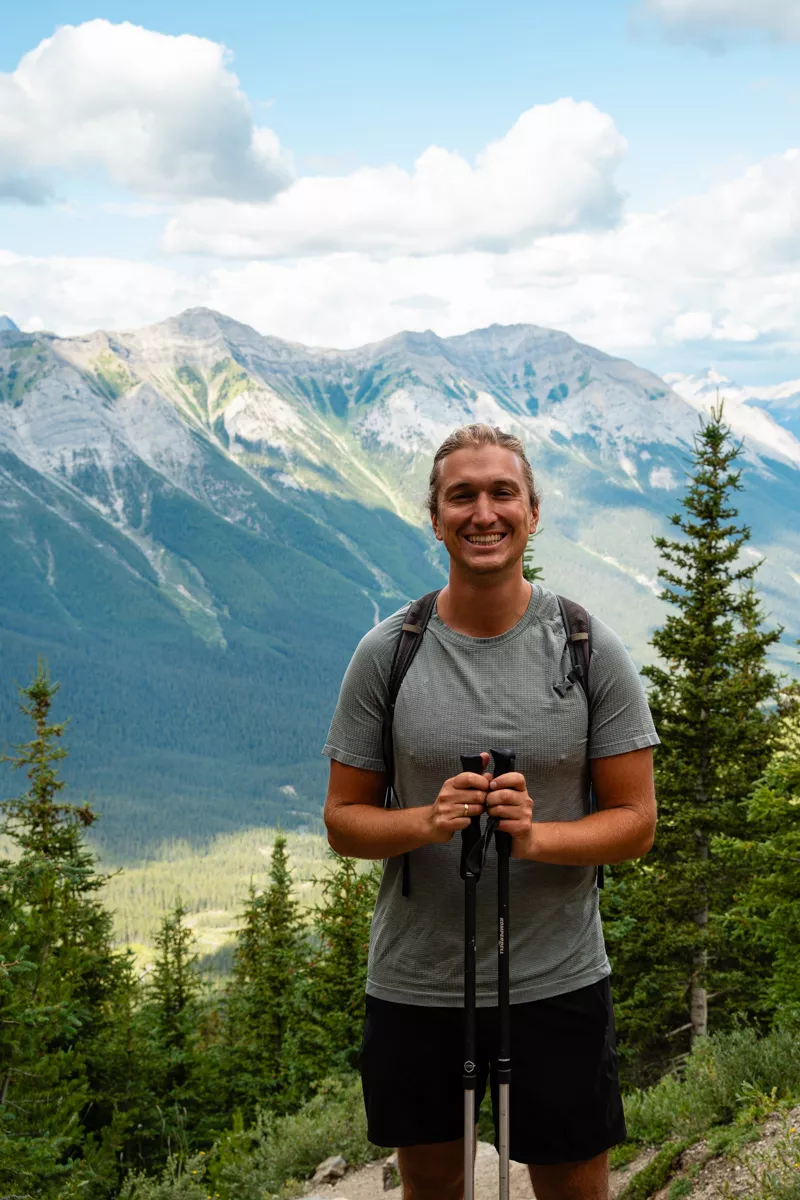 A smiling hiker poses with trekking poles in front of a stunning alpine backdrop, with forested valleys and jagged mountain peaks stretching behind him.