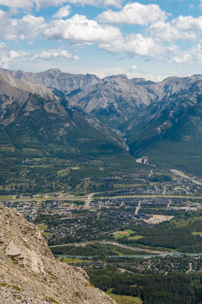 View from the summit of Ha Ling Peak overlooking the town of Canmore, with rows of houses, roads, and the Bow River visible far below. In the distance, rugged peaks of the Fairholme Range rise under a partly cloudy blue sky, creating a beautiful view across the Bow Valley.