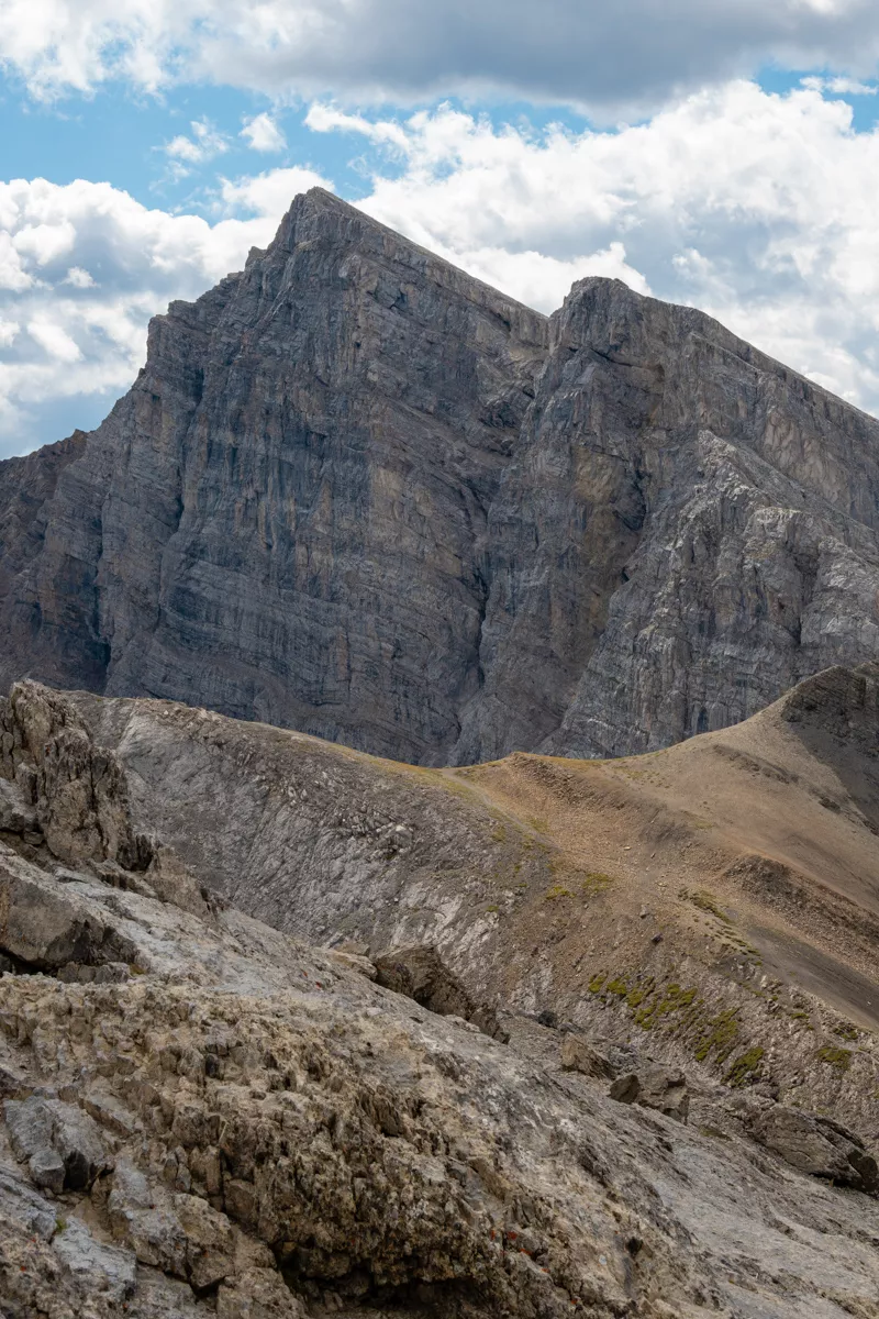 View from the summit of Ha Ling Peak looking toward a neighbouring mountain with steep, rugged cliffs and layered rock formations. The foreground shows part of the rocky summit ridge, highlighting the exposed alpine terrain under a partly cloudy sky.