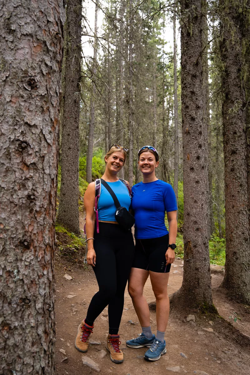 Two smiling hikers pose on a forest trail surrounded by tall pine trees. One wears a turquoise tank top with hiking boots and a backpack, and the other wears a blue athletic shirt with shorts and trail runners.