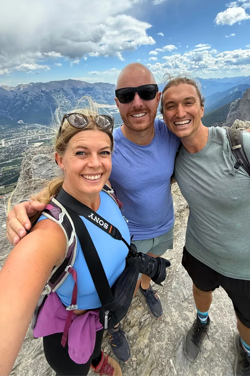 Three hikers smile for a selfie on the rocky summit of Ha Ling Peak, with a sweeping view of mountains and the town of Canmore in the background. The group is geared up with backpacks, hiking boots, and a camera, under a partly cloudy sky with dramatic light.