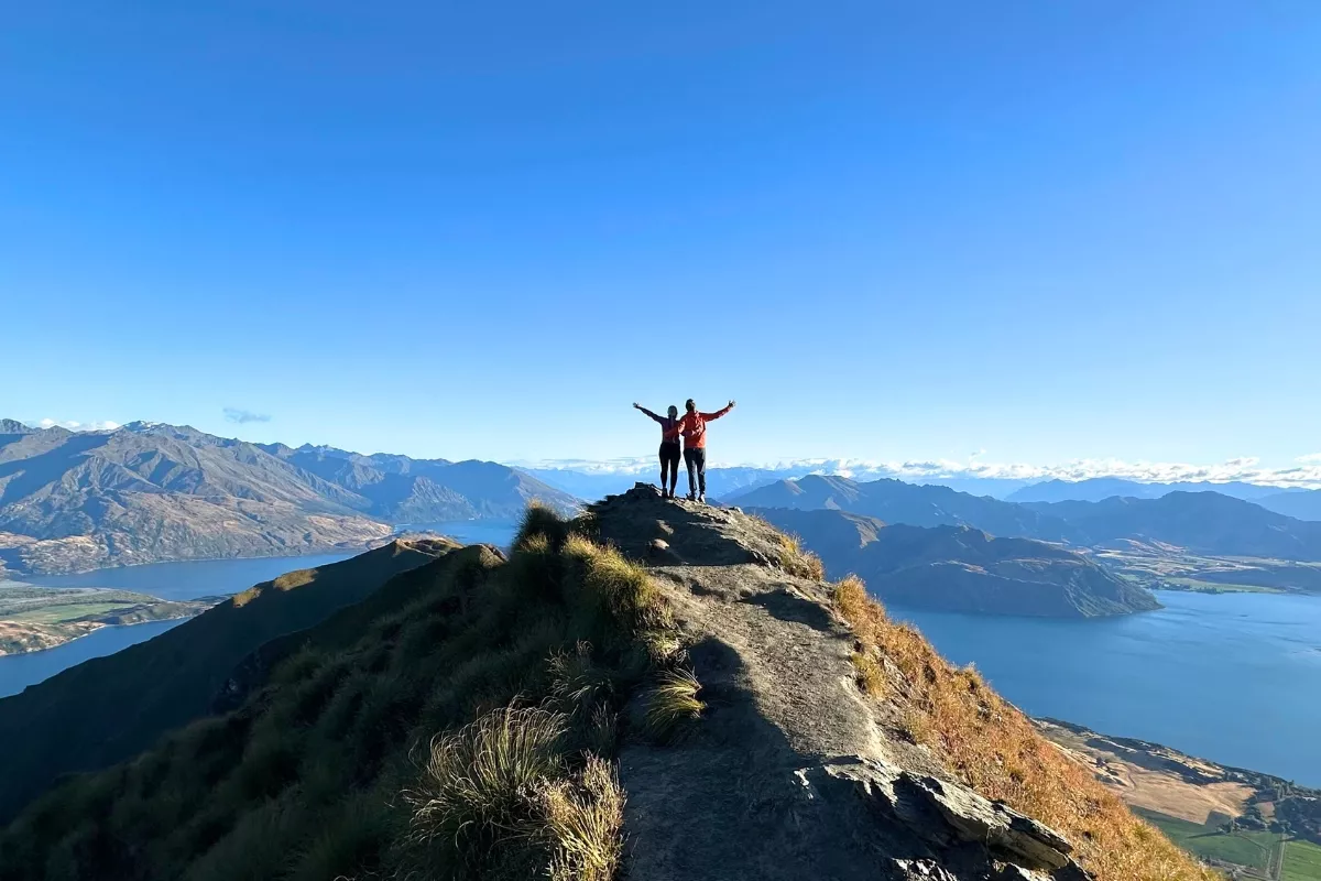 Two hikers stand with arms raised on the narrow ridgeline at Roys Peak, framed by a sweeping panoramic view of Lake Wanaka, mountain ranges, and valleys under a clear blue sky. The well-worn trail leads directly to the rocky viewpoint where they stand, emphasizing the dramatic elevation.