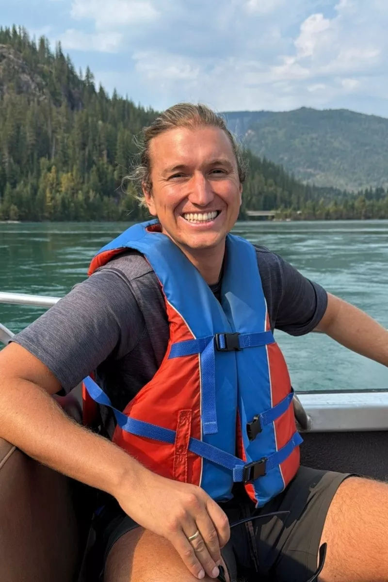 A man wearing a red and blue life jacket smiles while sitting on a boat, with forested mountains and calm river water in the background.