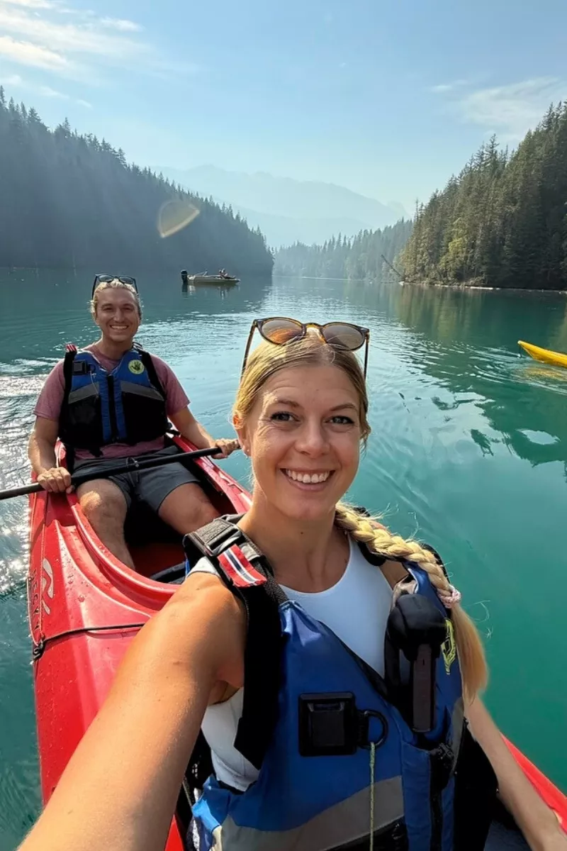 A woman takes a selfie from the front of a red tandem kayak while smiling with a man behind her, both wearing life jackets on a serene, tree-lined river. The couple paddles through calm, clear water with misty mountains visible in the distance.