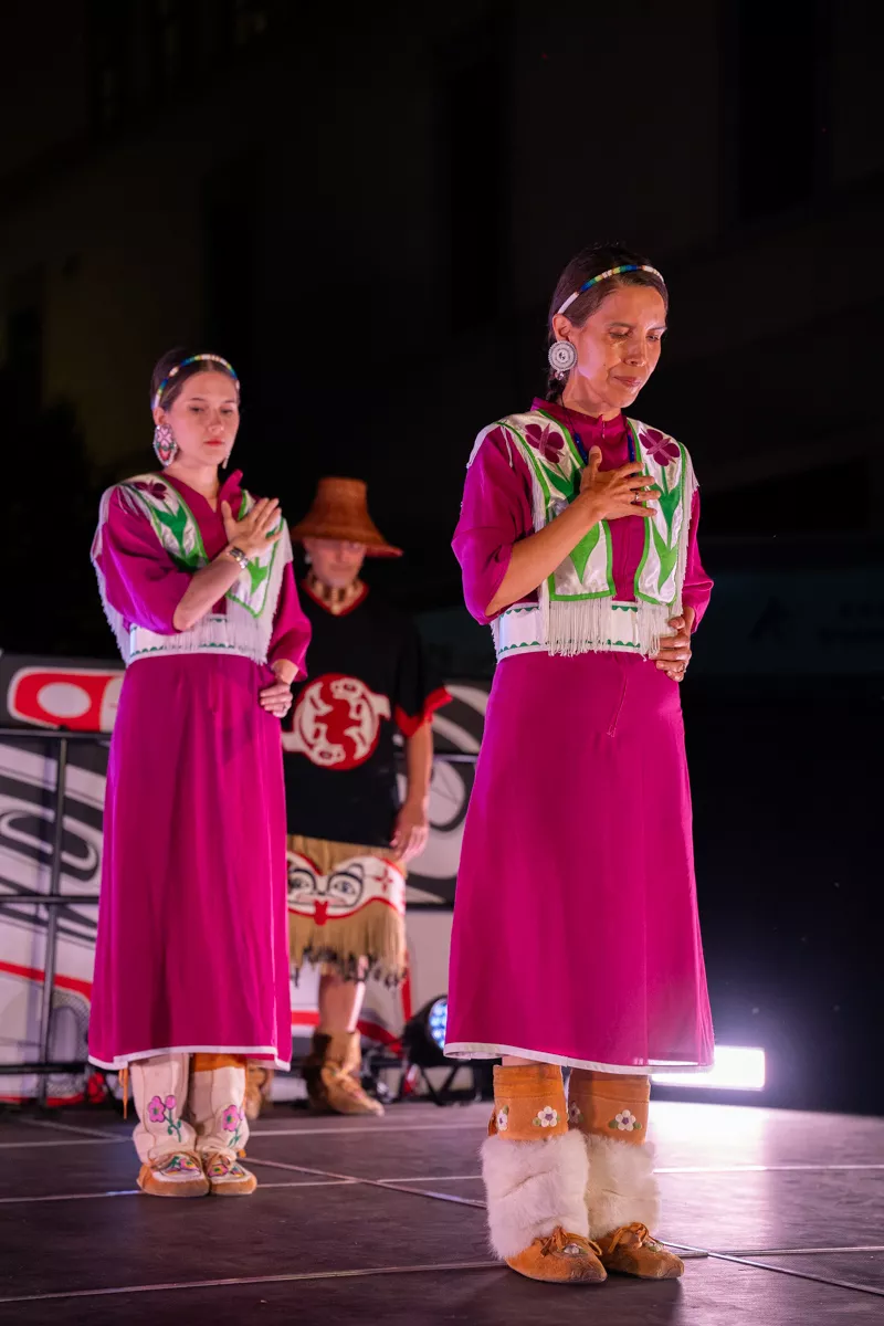 Two women in magenta dresses with beaded vests and fur-trimmed moccasins perform a traditional Indigenous dance on stage, with a man in regalia standing behind them. The performance is lit dramatically against a dark background during a cultural celebration.