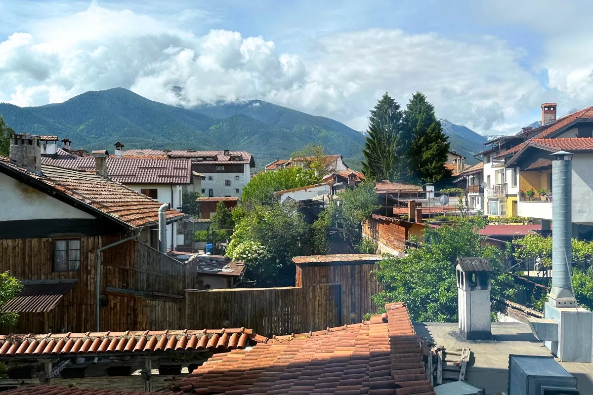 Red-roofed houses and rustic wooden buildings in Bansko, Bulgaria, with lush greenery and the Pirin Mountains in the background partially covered by clouds.