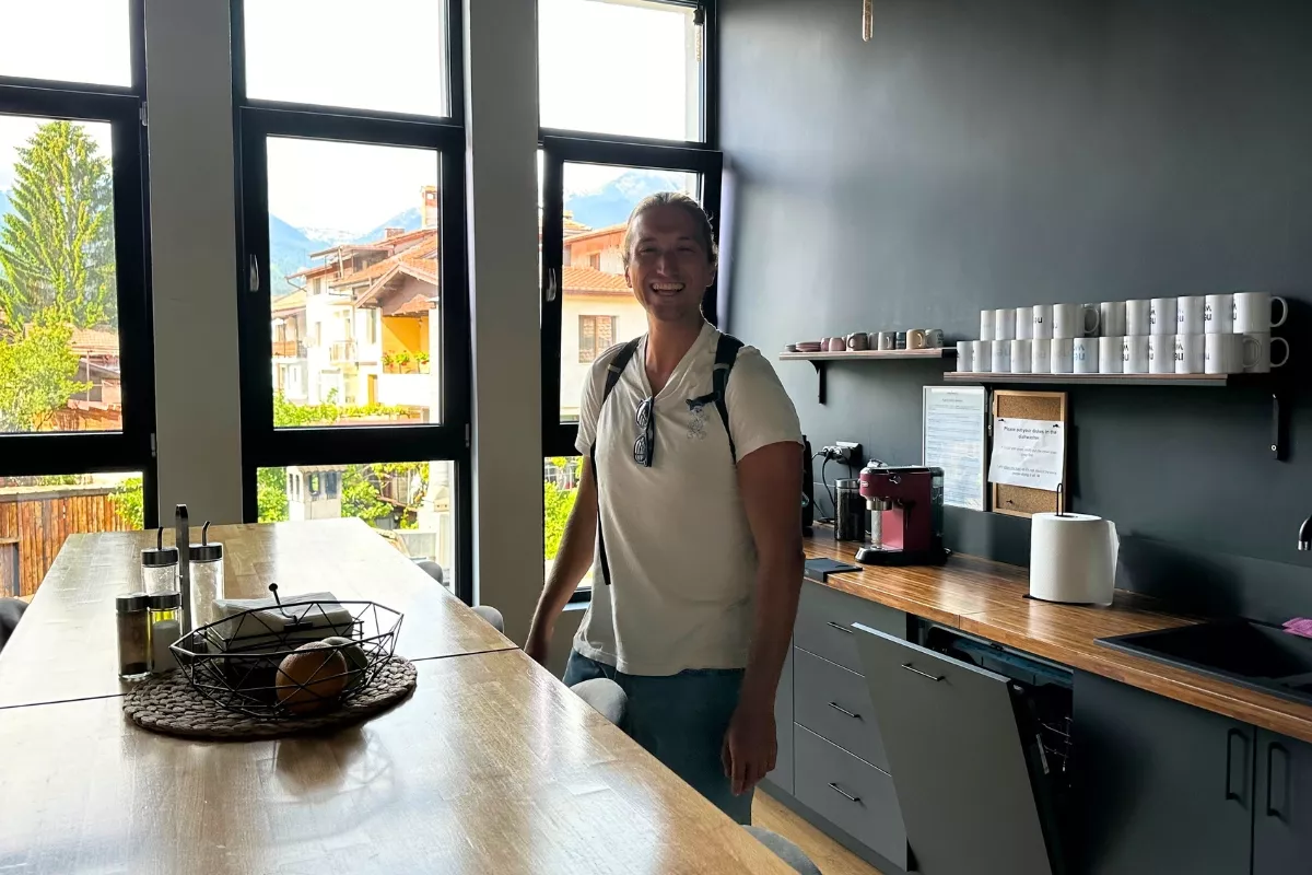 A man smiling inside a modern kitchen with black and wood finishes, standing near a window with a view of Bansko’s rooftops and the surrounding mountains.