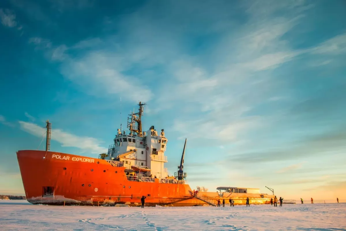 A large red icebreaker ship named Polar Explorer is stationed on a frozen sea at sunset. The sky is painted with soft blue and orange hues, while people in winter gear walk around the icy surface near the ship.