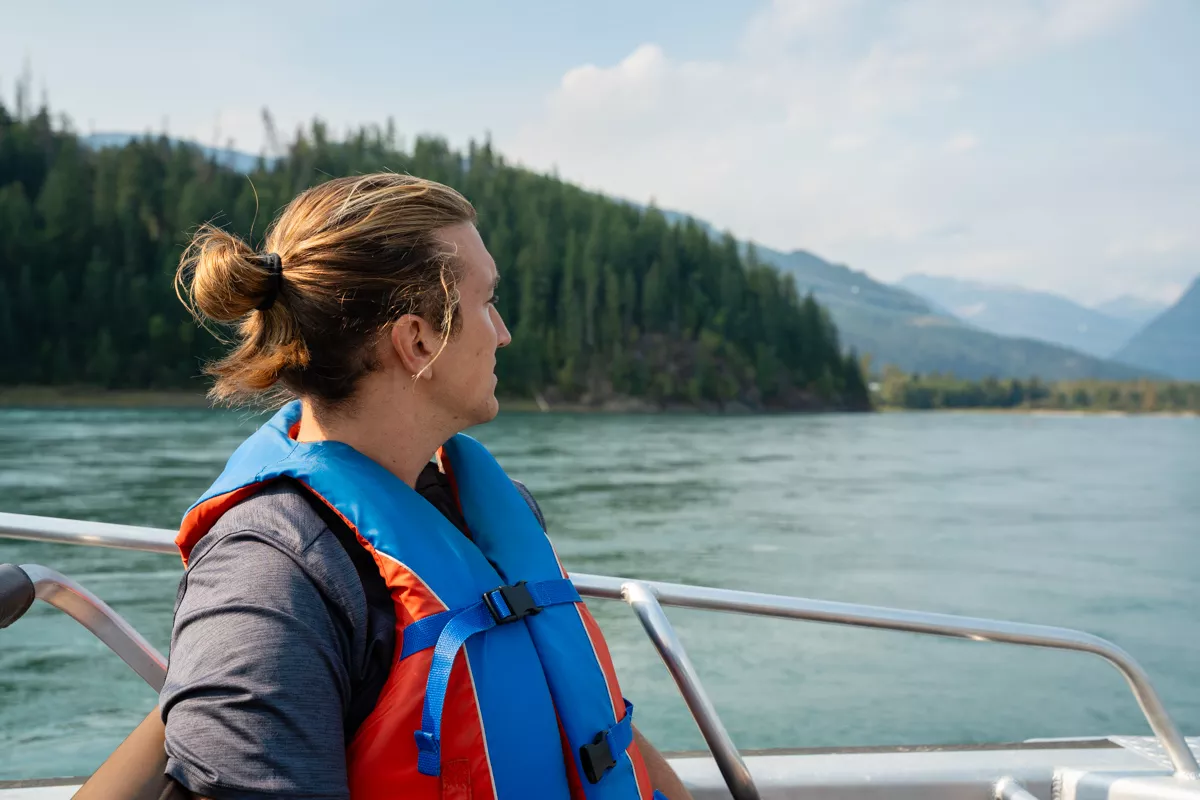 A man wearing a red and blue life jacket gazes out over a calm river surrounded by dense forest and distant mountains, seated on a boat with a metal railing. The peaceful scene captures a scenic outdoor adventure.