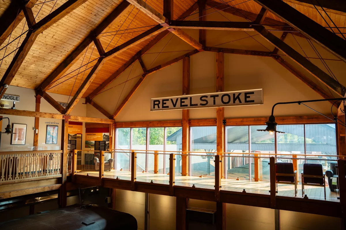 An interior view of a historic looking building with vaulted wooden ceilings and exposed beams, featuring a large “REVELSTOKE” railway sign mounted above a glass-railed walkway. Sunlight pours through tall windows, highlighting museum exhibits and vintage train artifacts below.