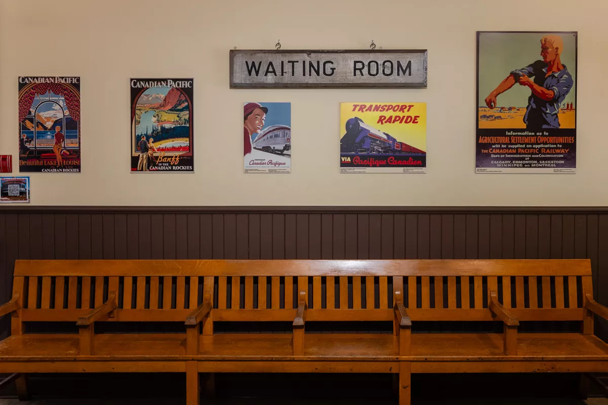 A vintage-style train station waiting room with a long wooden bench sits beneath a “WAITING ROOM” sign and a row of colorful retro Canadian railway posters. The posters advertise scenic travel through the Canadian Rockies and historic immigration opportunities via the Canadian Pacific Railway.
