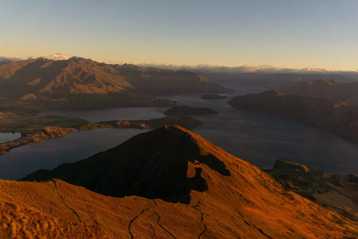 A wide view from Roys Peak at sunrise, showing the winding hiking trail along the golden-lit ridgeline with Lake Wānaka and its islands below. The surrounding mountains are partially cast in shadow with soft morning light hitting the peaks.