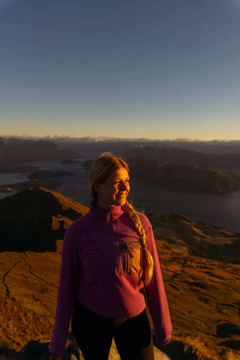 A woman in a pink fleece stands smiling on Roy's Peak at sunrise, with golden light on her face and Lake Wānaka along with the distant ranges of Mount Aspiring National Park behind her.