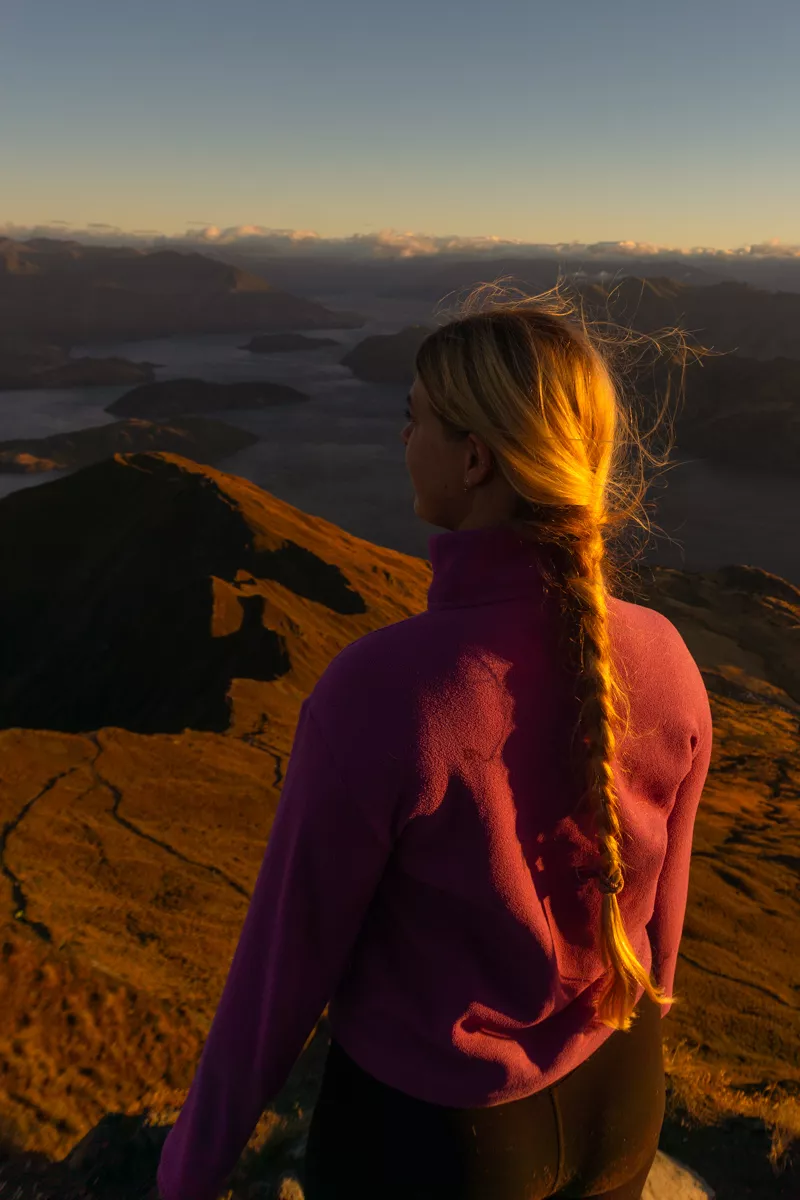 A woman with a long braid stands facing the sunrise from Roys Peak, overlooking Lake Wanaka and the mountain ridges glowing in early morning light.