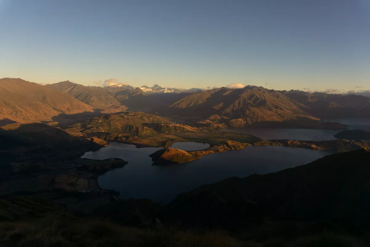 A panoramic view from Roys Peak at sunrise, showing Lake Wanaka and its scattered islands with the surrounding mountains catching the early morning light. Snow-capped peaks are visible in the distance under a clear sky.