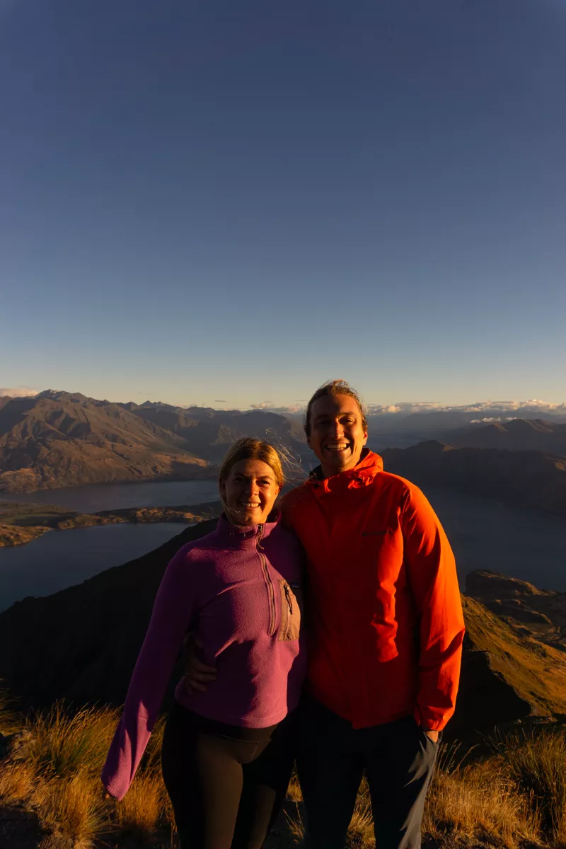 A smiling couple stands on a mountain at sunrise, dressed in bright hiking gear with a stunning view of Lake Wanaka and surrounding peaks behind them, bathed in golden morning light.