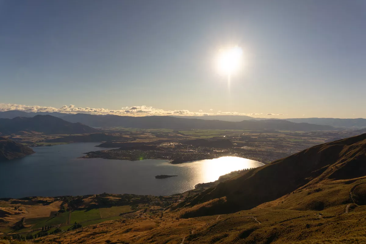 A bright morning sun reflects off Lake Wanaka as seen from Roys Peak, casting a strong glare on the water below. The town of Wanaka and surrounding farmland stretch across the valley, with rolling hills and mountains in the distance.