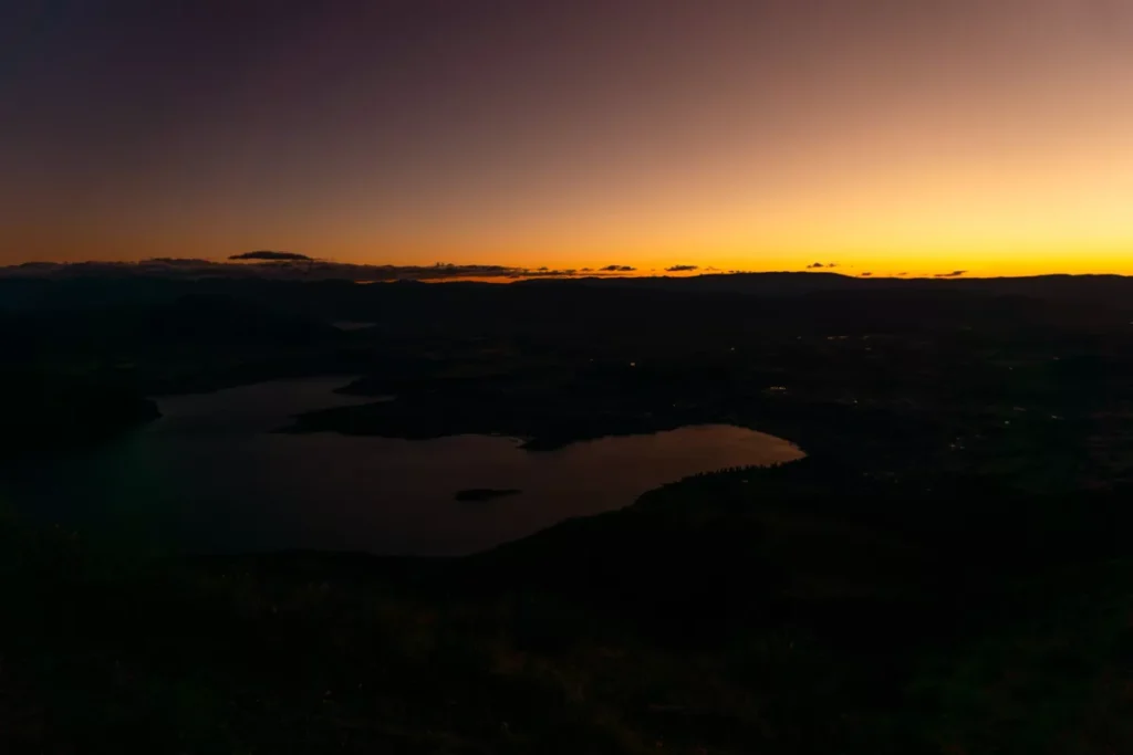 A pre-dawn view over Lake Wānaka from Roys Peak, with the sky glowing in deep orange and purple hues above the dark mountain silhouettes. The lake and landscape below are dimly lit, capturing the stillness just before sunrise.
