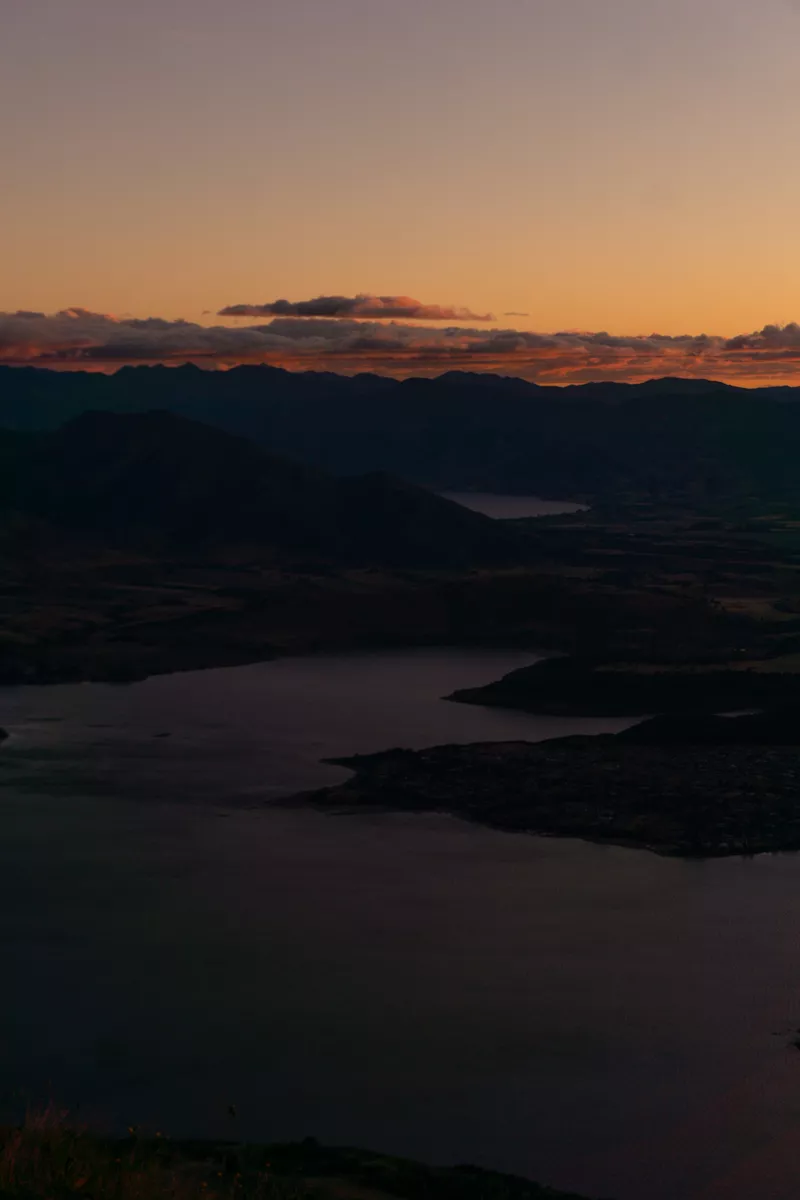 A view over Lake Wanaka at dawn, with the town just visible in the foreground and silhouetted mountains lining the horizon. The sky glows in soft orange and purple tones, with scattered clouds catching the early morning light.