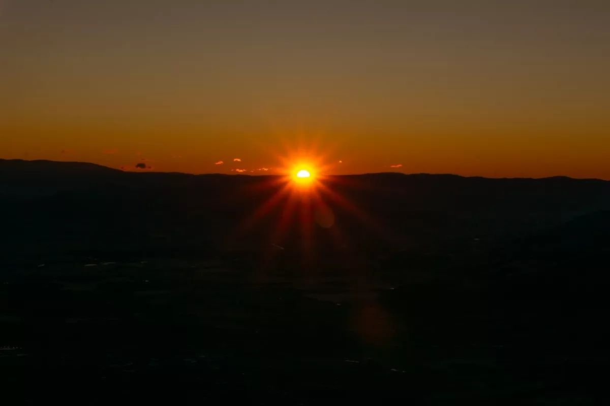 The sun rises just above the dark mountain horizon, casting golden-orange rays across a clear sky. The landscape below remains in shadow, creating a dramatic contrast between light and dark in this early morning view.