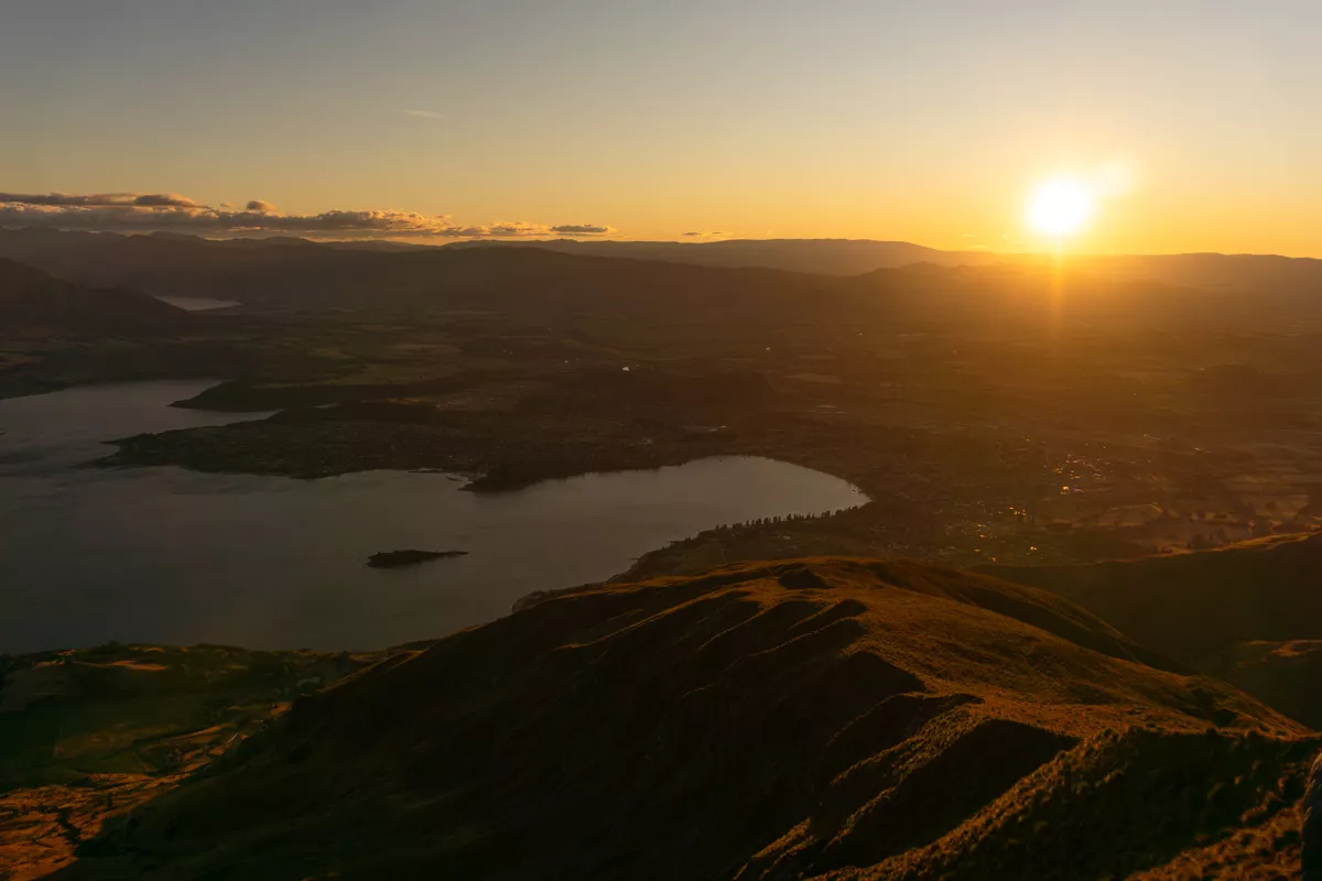Sunrise view from Roy's Peak overlooking Lake Wānaka, with golden sunlight illuminating the ridgeline in the foreground and casting a warm glow across the town and surrounding hills below. The sun sits just above the horizon in a clear morning sky.