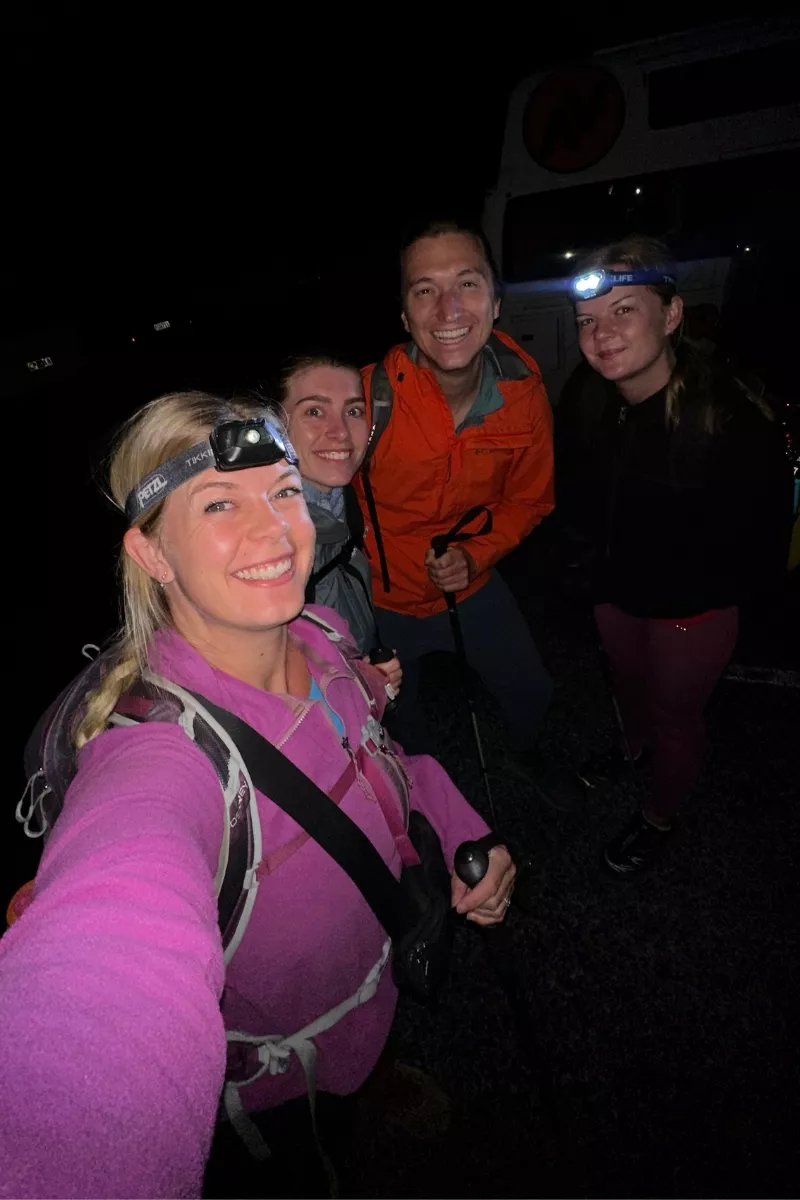 Four hikers wearing headlamps and outdoor gear smile for a selfie in the dark, standing on a gravel surface with faint outlines of vehicles behind them.