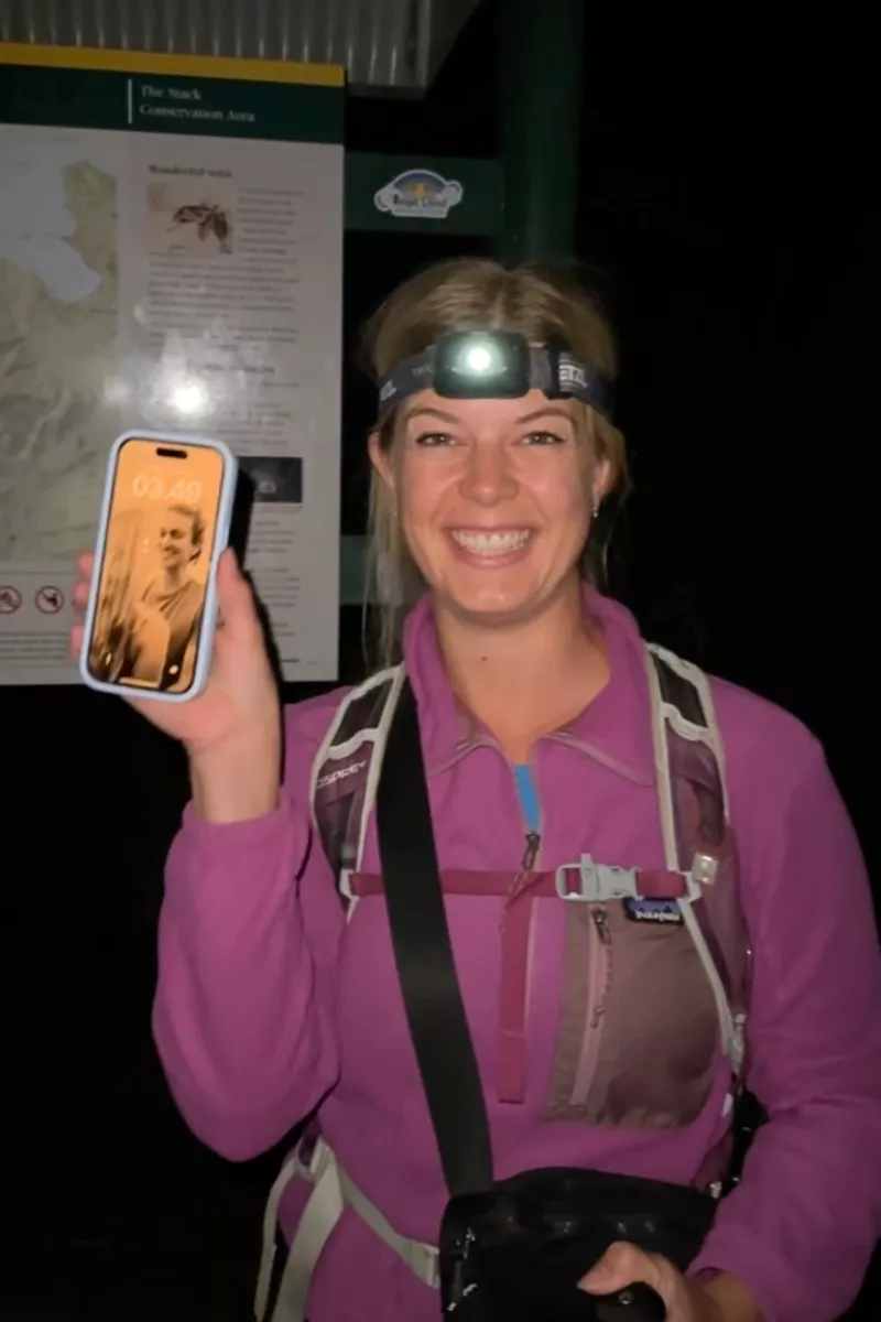A smiling hiker wearing a headlamp and purple jacket holds up a phone showing the time 03:49 AM, standing in front of a trailhead sign for “The Neck Conservation Area” in the dark. Her backpack and hiking poles suggest an early start to a trek.