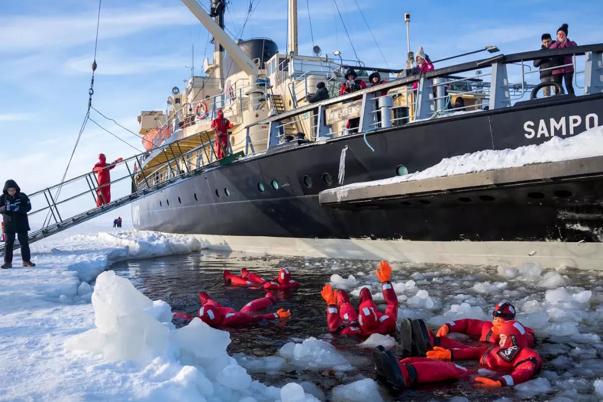 People in red survival suits float in icy water near a large black and cream icebreaker ship named "SAMPO," which is docked in a frozen landscape. The ship is surrounded by snow and ice.